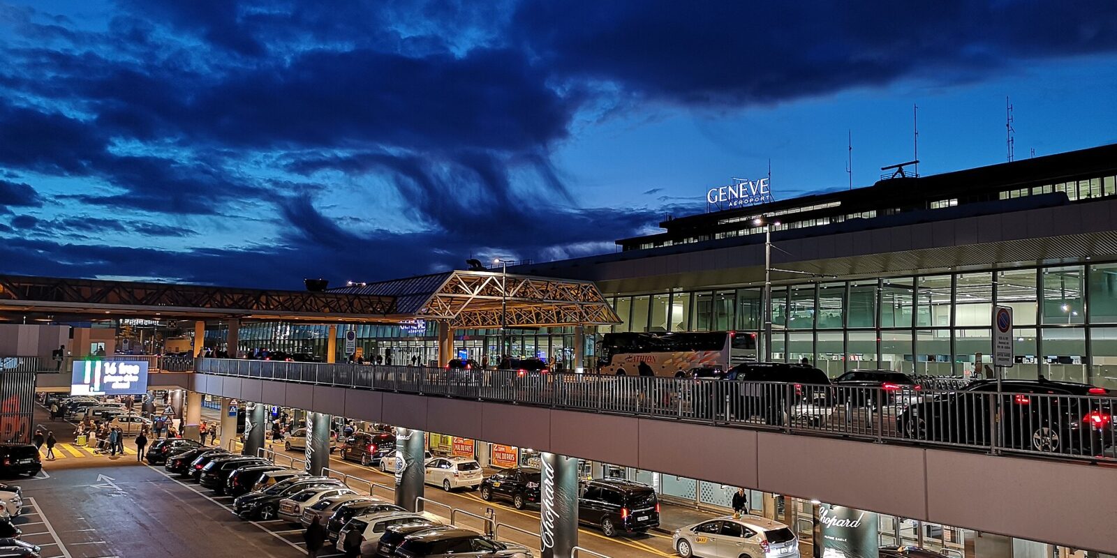 Geneva Airport terminal frontage lit at dusk with taxis and a coach at the kerbside