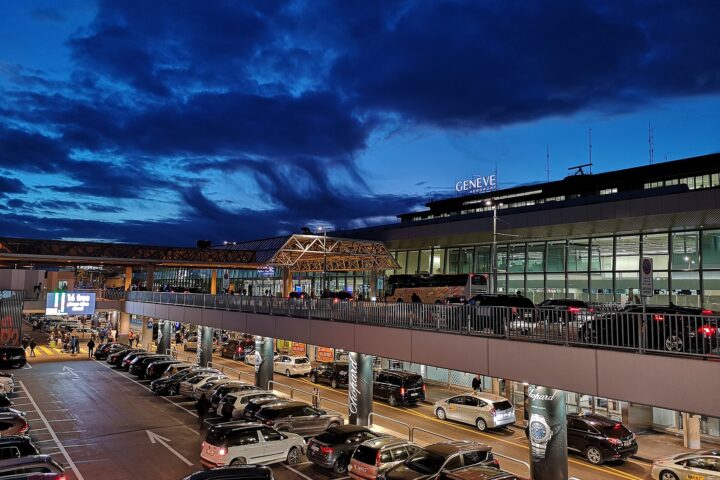 Geneva Airport terminal frontage lit at dusk with taxis and a coach at the kerbside