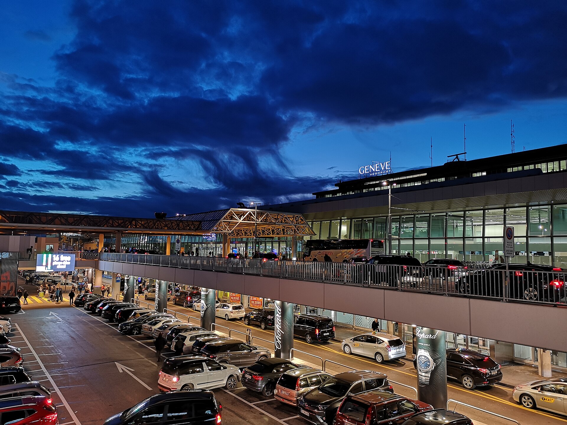 Geneva Airport terminal frontage lit at dusk with taxis and a coach at the kerbside