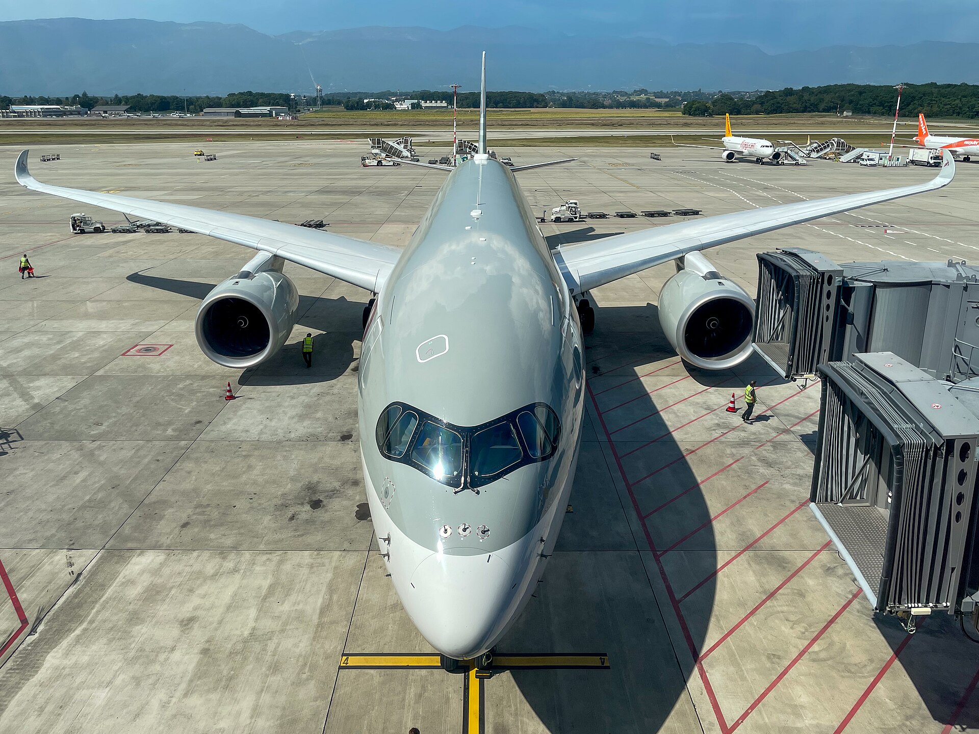 A wide-body jet parked at a Geneva Airport gate with the Jura mountains beyond the apron