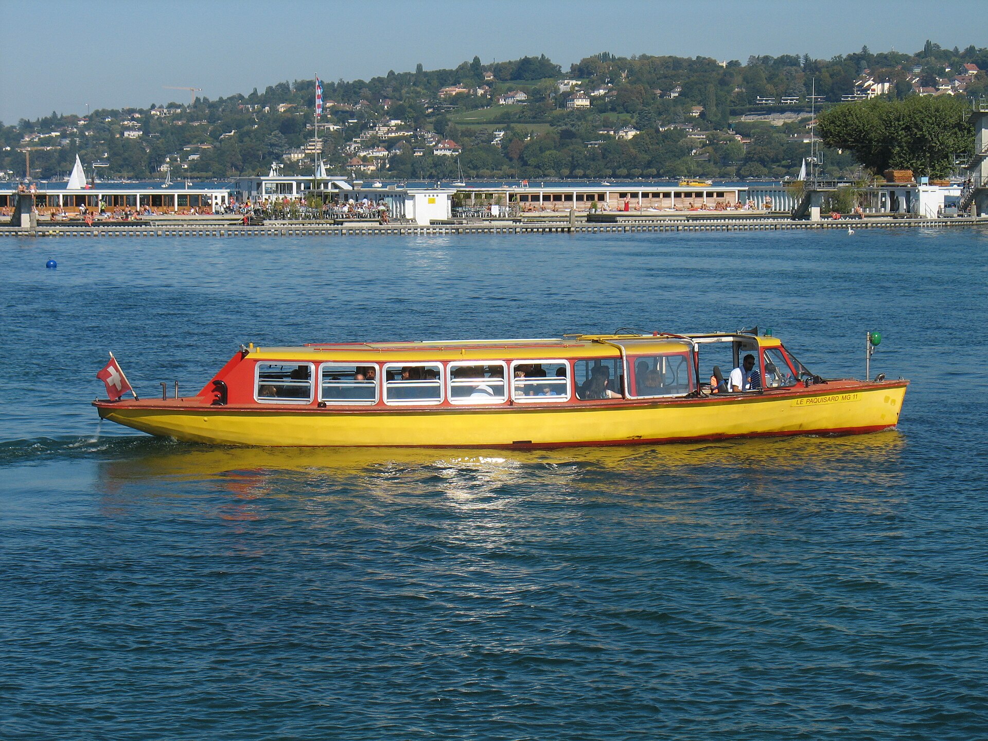 A yellow Mouettes Genevoises water taxi crossing Geneva harbour in front of the Bains des Pâquis