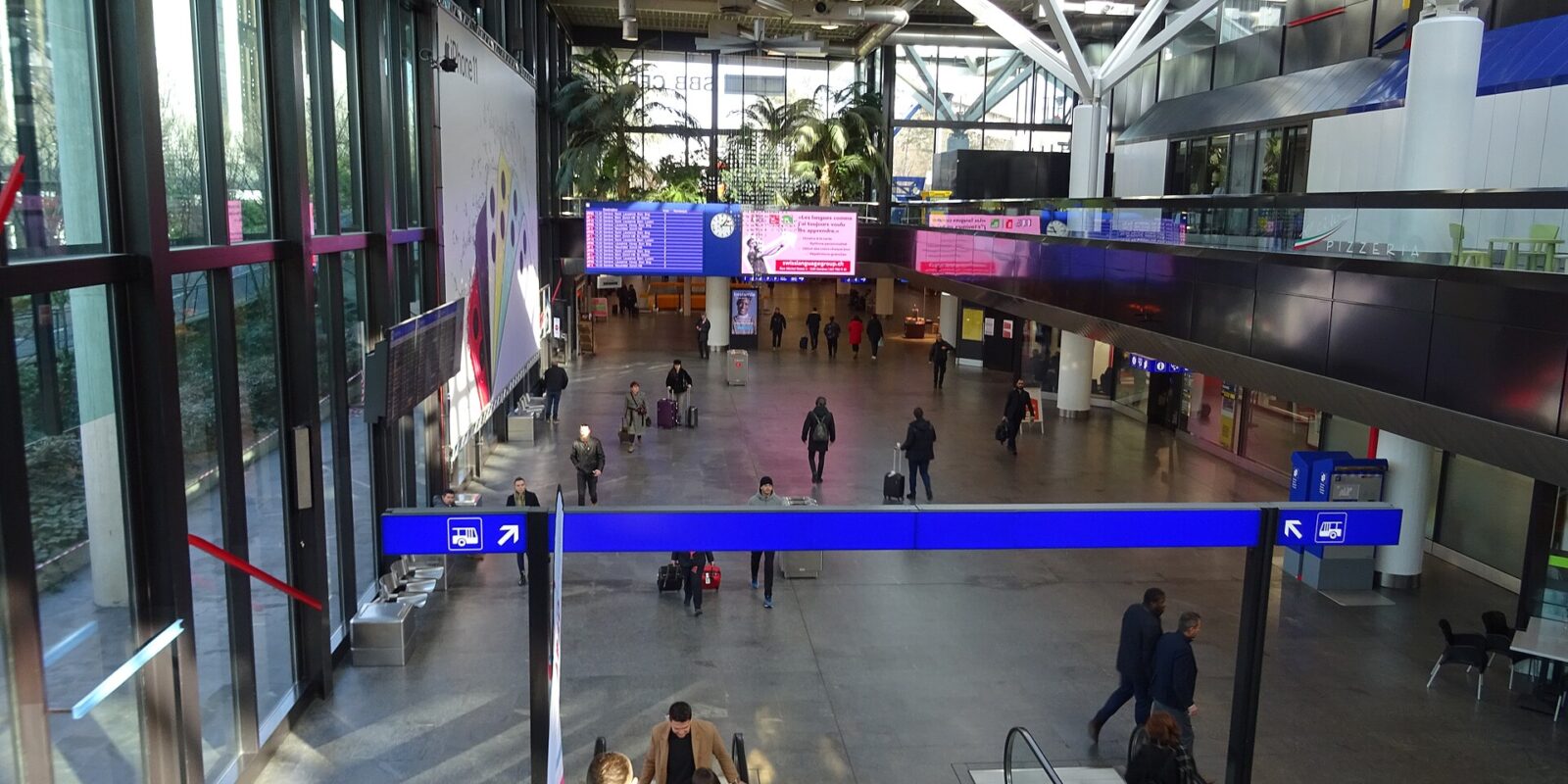 The main hall at the Genève-Aéroport station, with escalators, a departure board and the SBB sign behind the palms