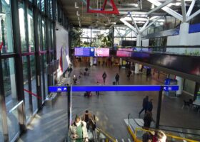 The main hall at the Genève-Aéroport station, with escalators, a departure board and the SBB sign behind the palms