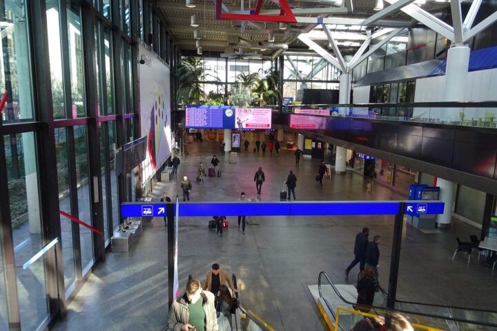 The main hall at the Genève-Aéroport station, with escalators, a departure board and the SBB sign behind the palms