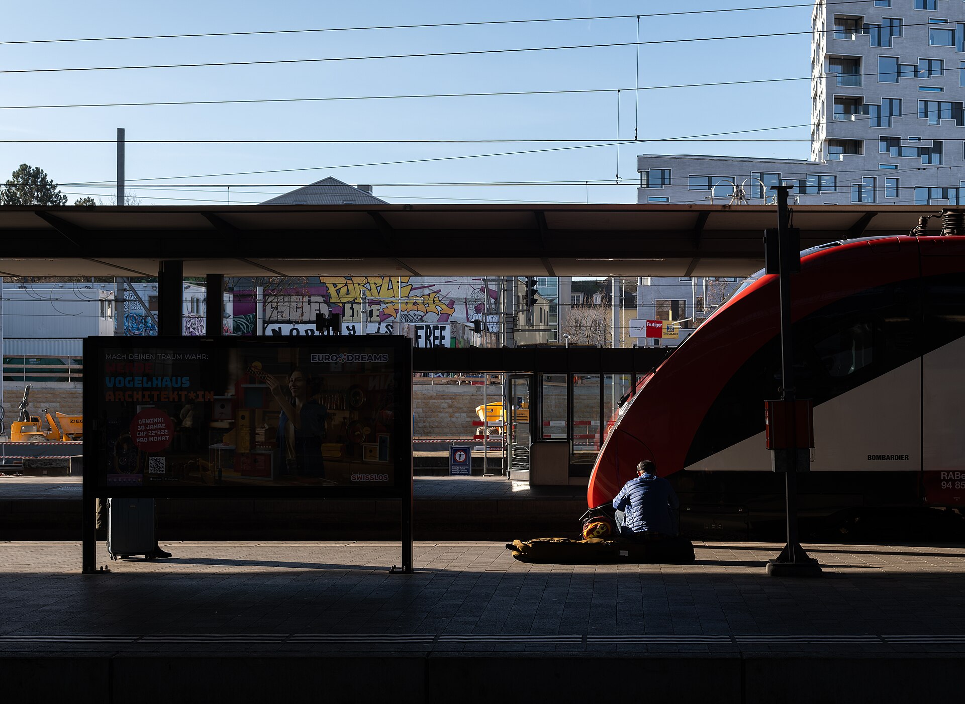 A traveller sitting beside a snowboard bag on a Swiss railway platform next to a red regional train