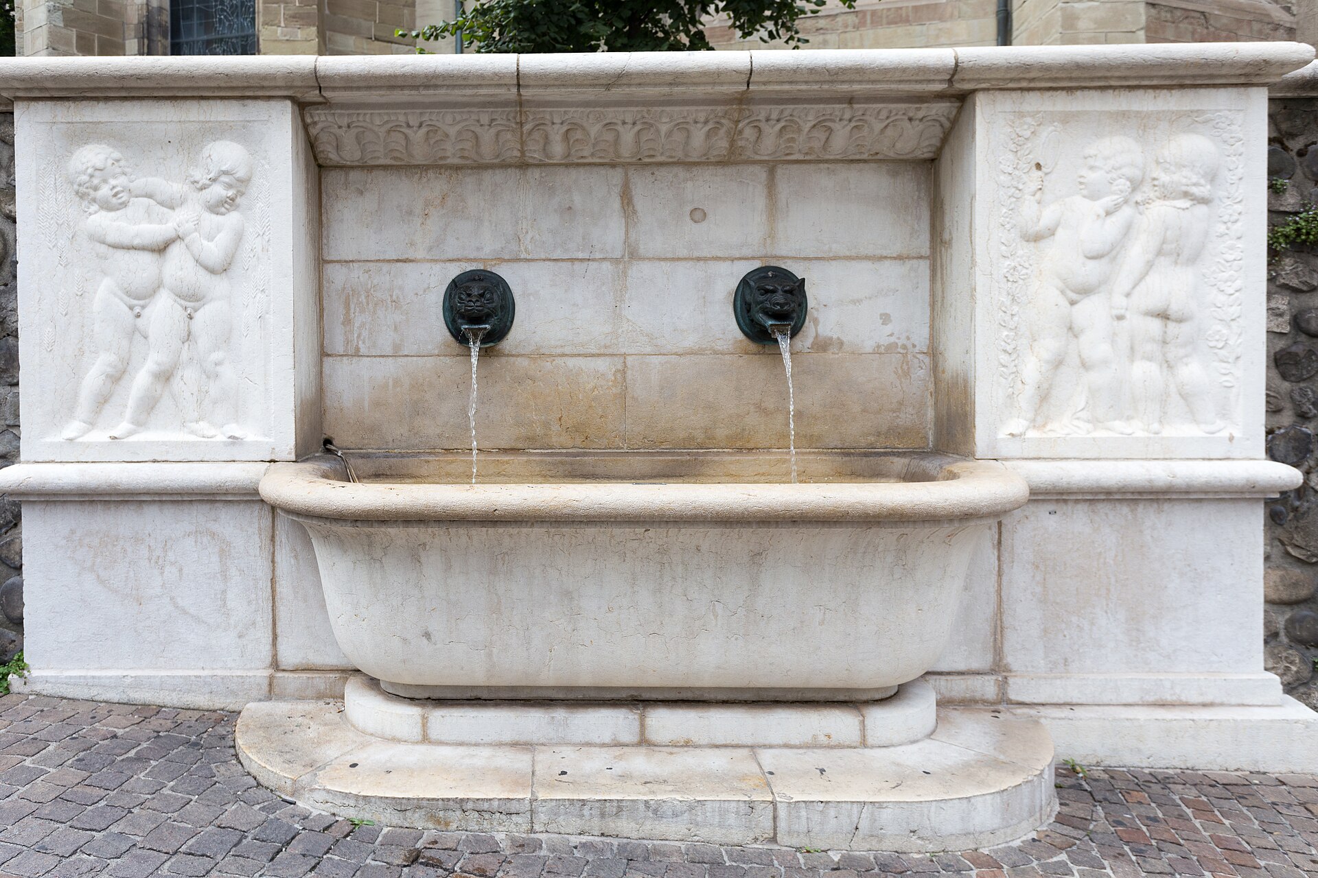 A white stone public fountain in Geneva with two bronze spouts and carved reliefs of playing children