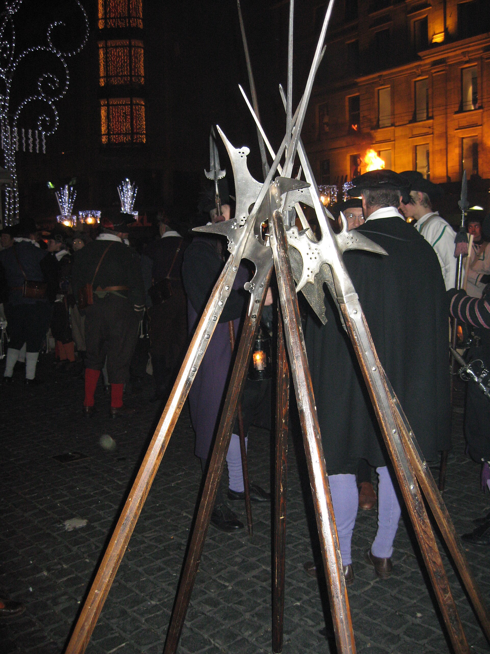 Halberds stacked in a tripod on a cobbled street at night during the Escalade, with costumed participants and Christmas lights