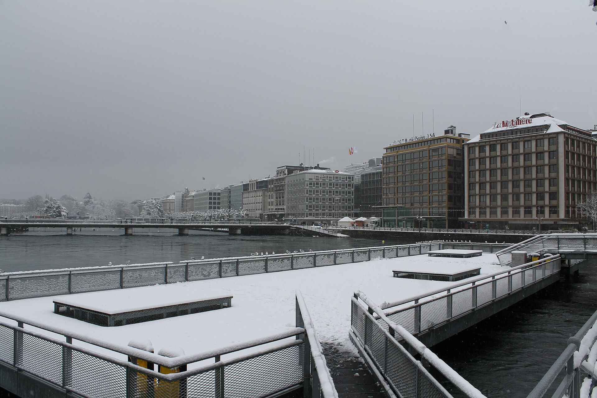 A snow-covered waterfront deck on the Rhône in central Geneva under a grey winter sky