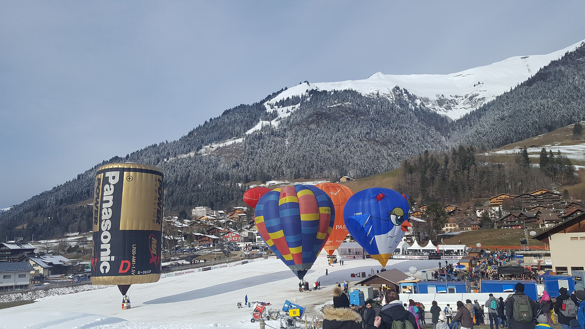 Hot-air balloons, including a giant battery and a bird, inflating on a snowy field at Château-d'Œx