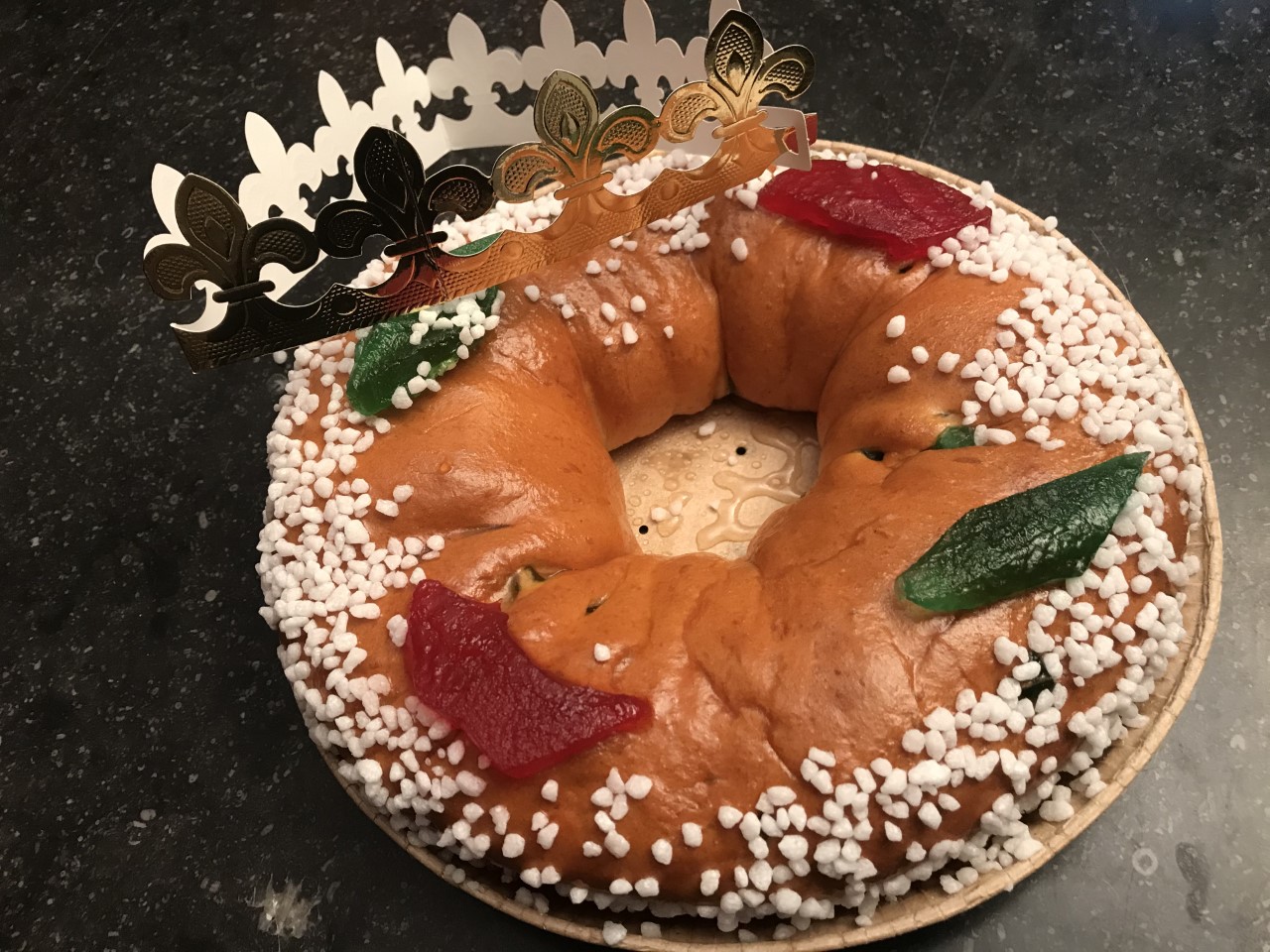 A brioche crown cake for Epiphany topped with pearl sugar and candied fruit, with a gold paper crown