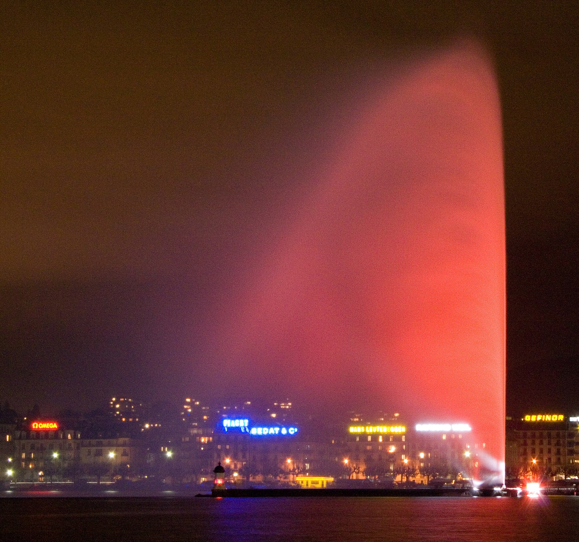 The Jet d'Eau illuminated red and pink at night against the lights of Geneva's lakefront