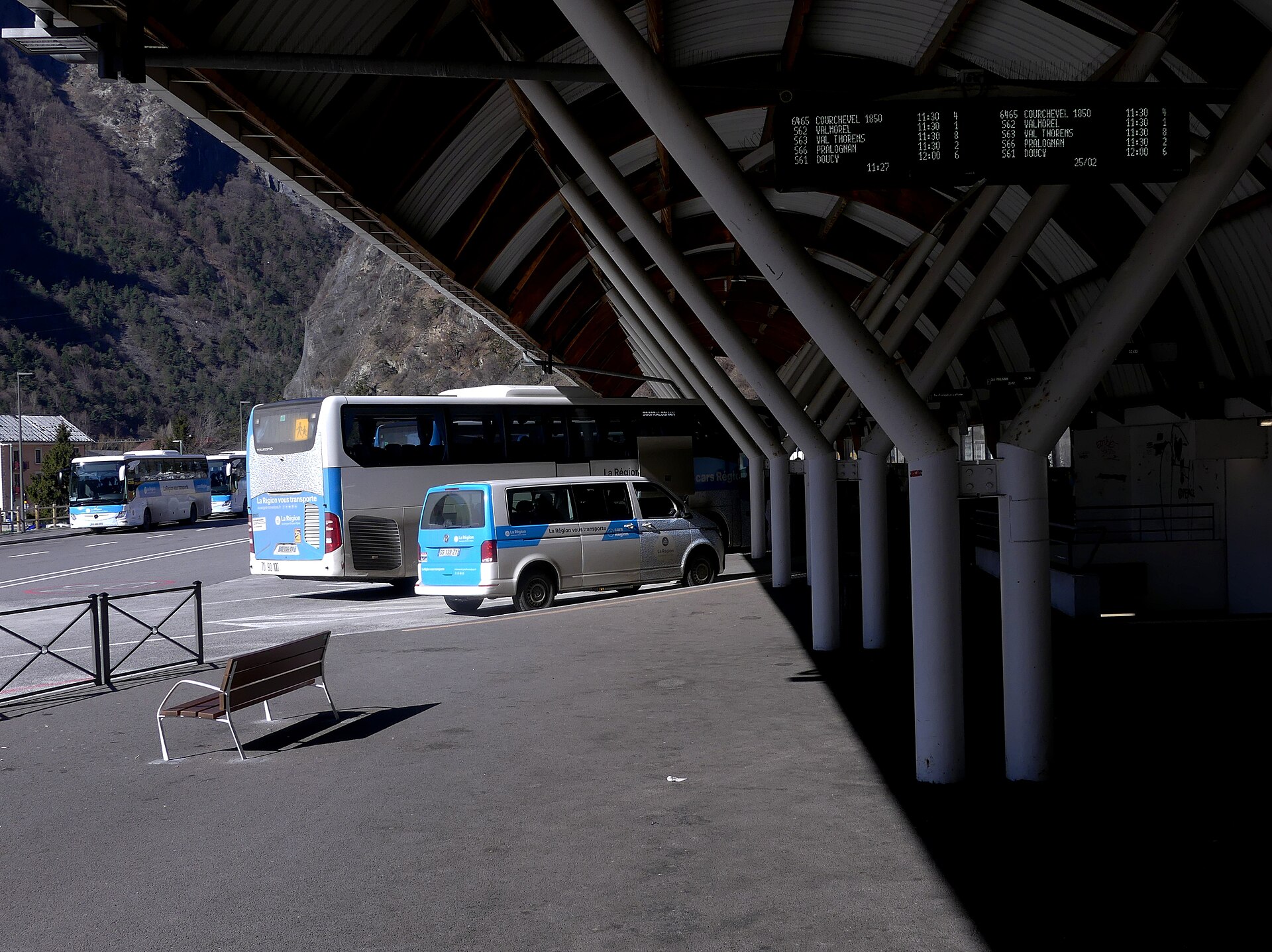 Coaches and a minibus under the canopy at Moutiers bus station with a departure board listing ski resorts