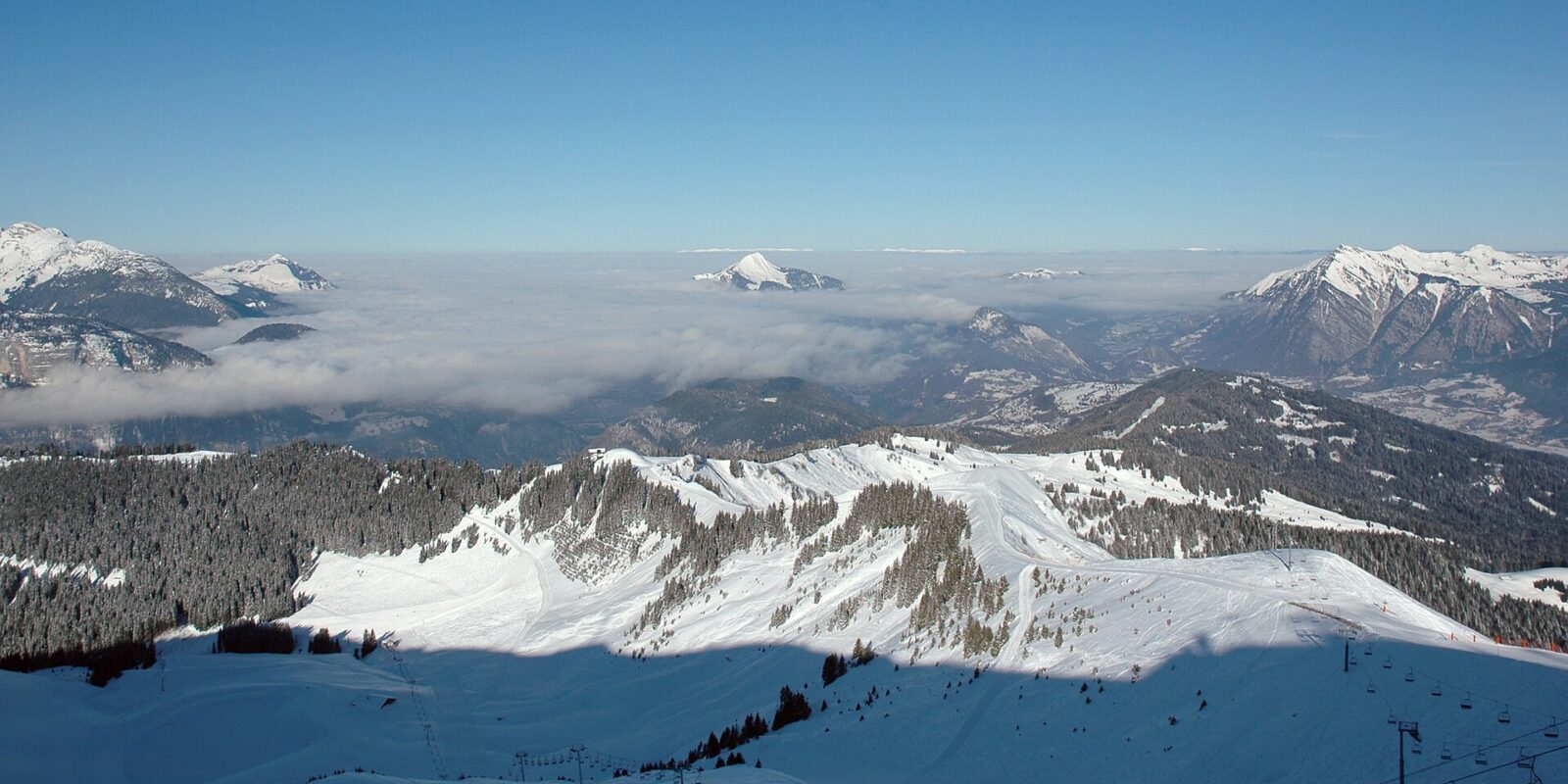 Snow-covered pistes and forested ridges of the Grand Massif above a cloud inversion filling the Arve valley