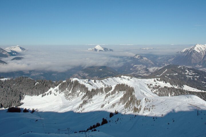 Snow-covered pistes and forested ridges of the Grand Massif above a cloud inversion filling the Arve valley