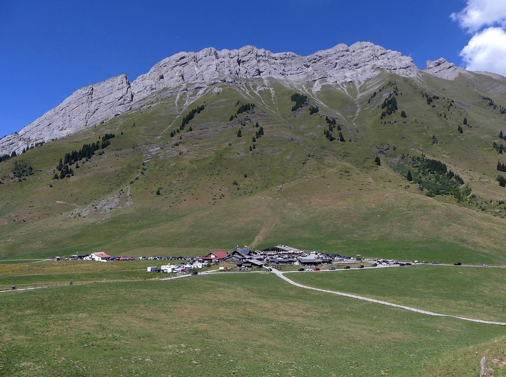 The limestone wall of the Aravis chain rising above the Col des Aravis, with chalets and a car park on the pass below