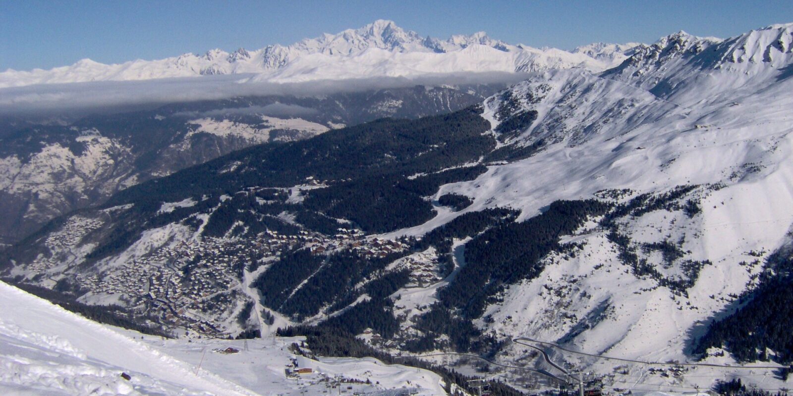 The Méribel valley under snow seen from the pistes above, with the resort's neighbourhoods on the hillside and Mont Blanc on the horizon