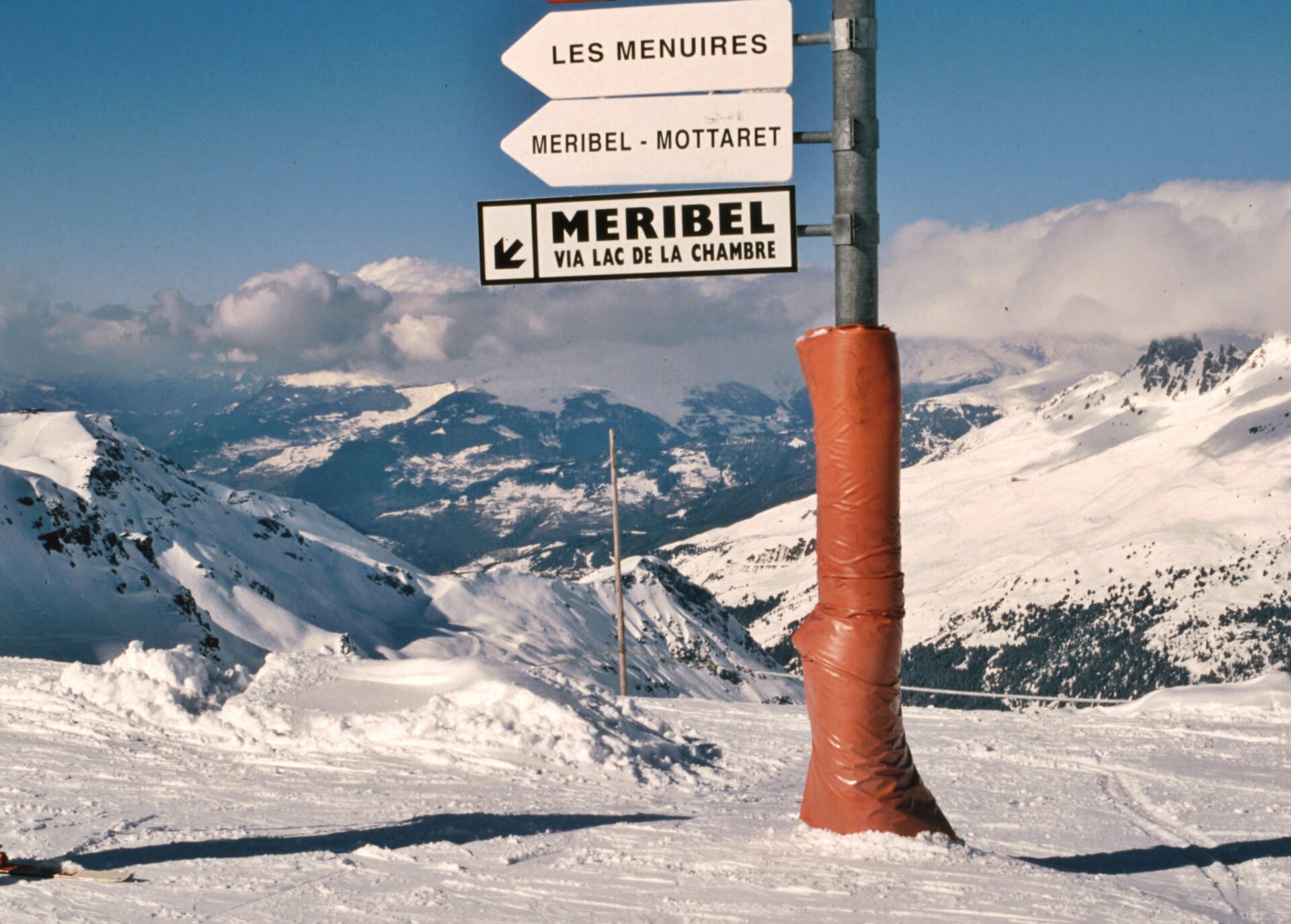 A piste signpost on a snowy col pointing to Les Menuires, Méribel-Mottaret and Méribel via Lac de la Chambre