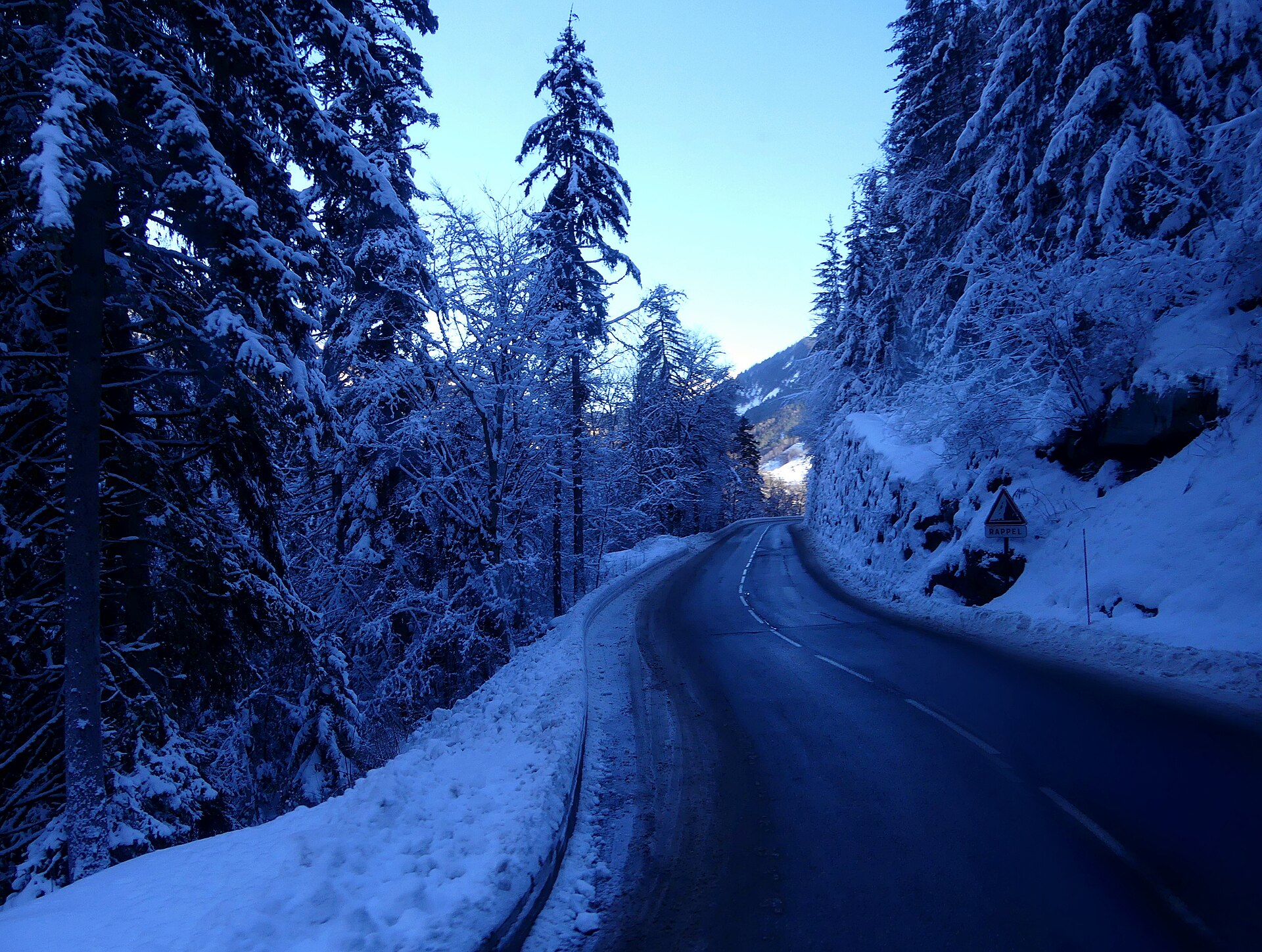 A cleared but snow-lined mountain road through pine forest in the Belleville valley at dusk in December