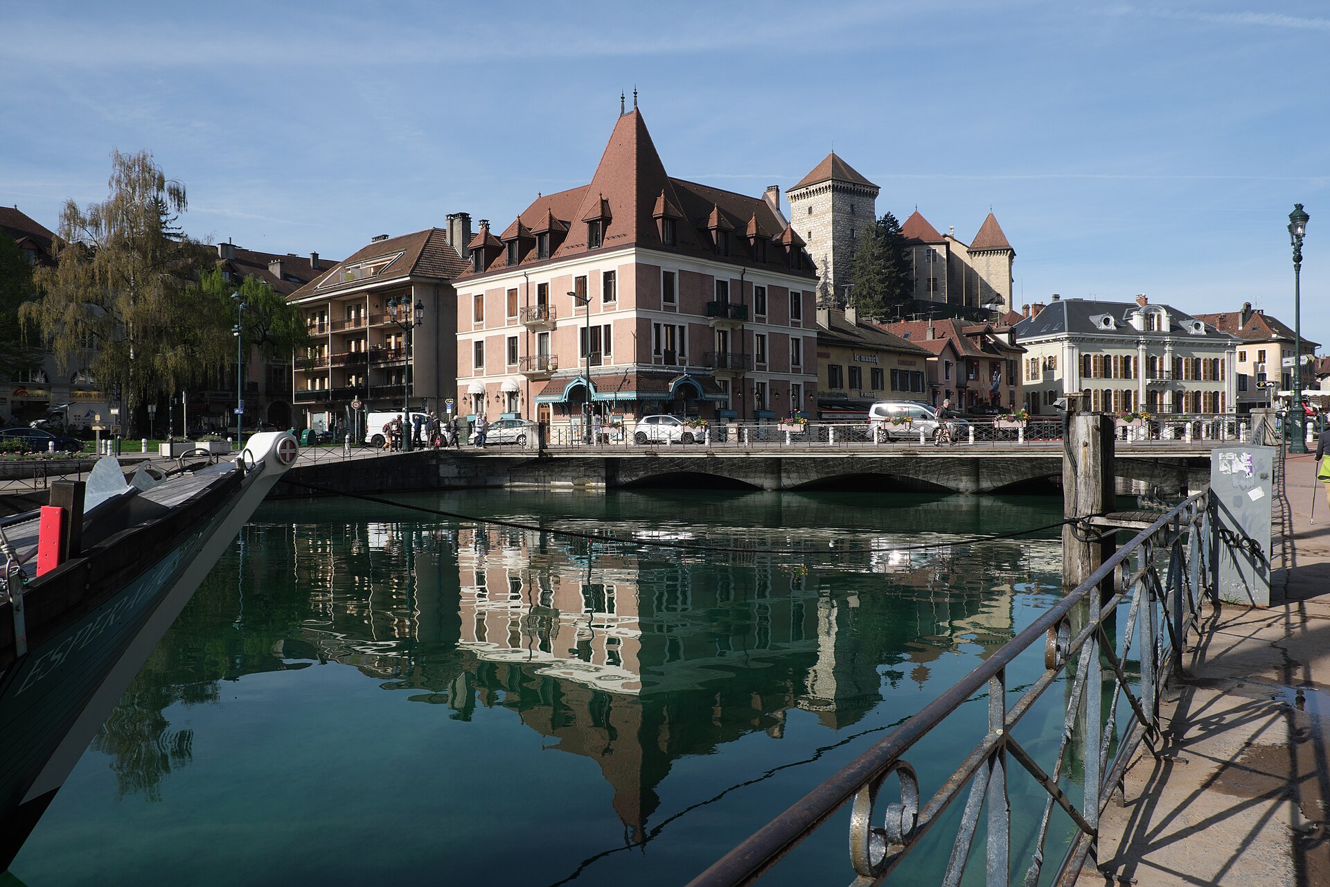 Annecy castle rises behind old-town roofs above a bridge, with the buildings reflected in the green canal below.