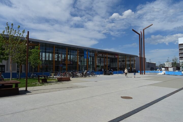 Annemasse railway station's glass facade faces a broad paved forecourt with parked bicycles and pedestrians beneath a cloudy blue sky.