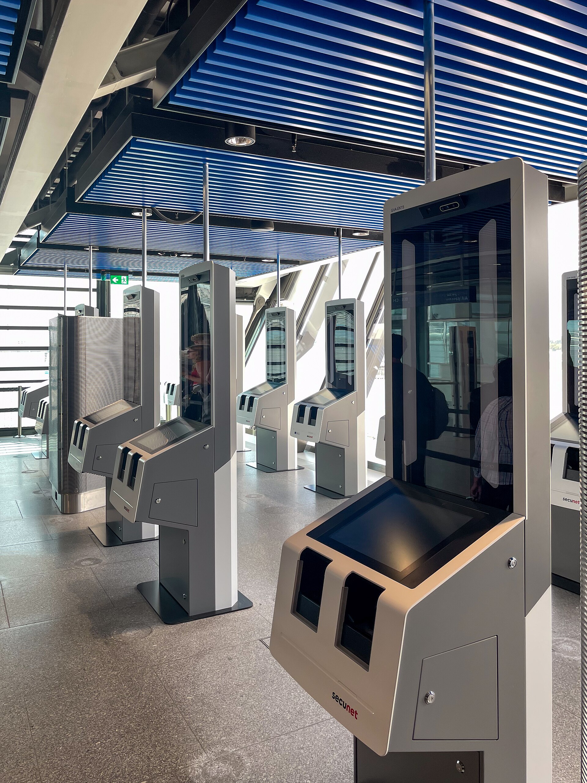 Automated passport gates inside Geneva Airport have document readers and tall camera screens beneath a blue slatted ceiling.