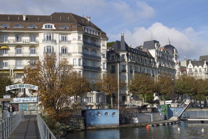 Autumn trees line Geneva's waterfront beneath ornate hotel façades, viewed from the Bains des Pâquis jetty.