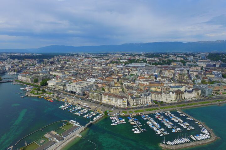 Bains des Pâquis bathing enclosures extend into Lake Geneva beside the marina, with waterfront buildings and mountains beyond.