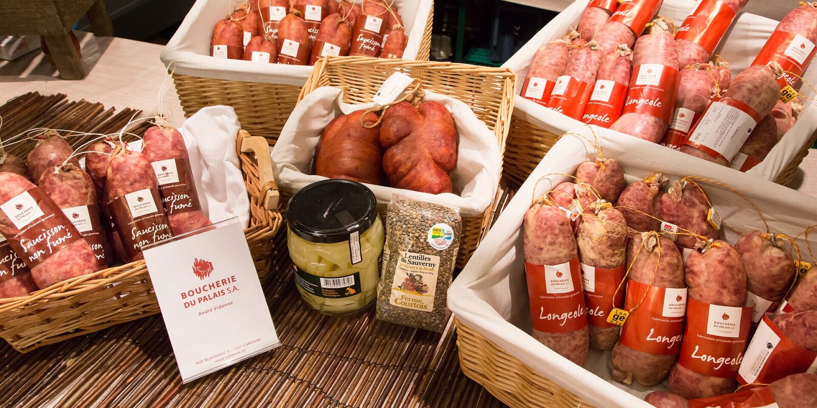 Baskets of longeole sausages with red labels reading Longeole and artisanal Geneva production, next to smoked saucissons, a jar of cardoons and a bag of lentils