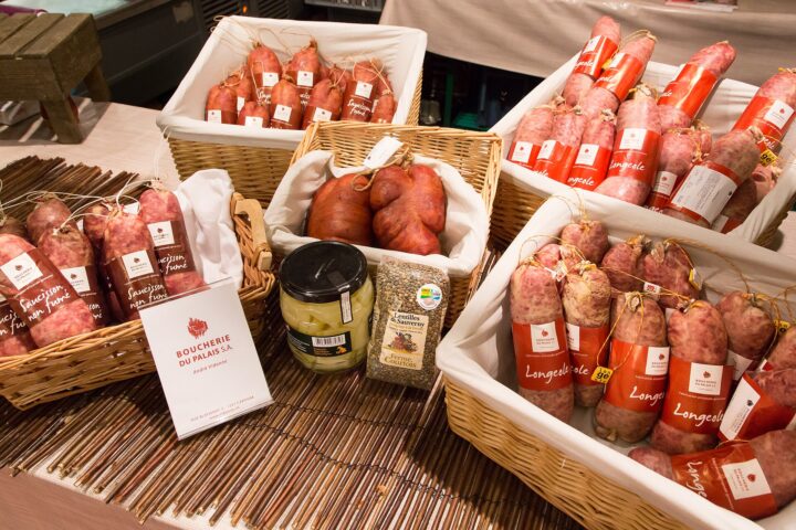 Baskets of longeole sausages with red labels reading Longeole and artisanal Geneva production, next to smoked saucissons, a jar of cardoons and a bag of lentils