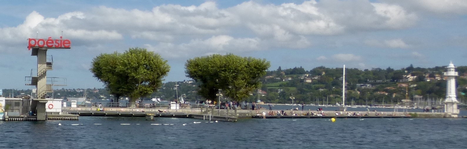 Bathers relax beside Lake Geneva at the Bains des Pâquis, with the diving tower, leafy trees and lighthouse framing the bathing platforms.