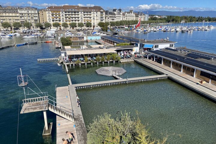Bathers stand beside the lake basin at Geneva's Bains des Pâquis, with diving platforms and waterfront hotels beyond the water.