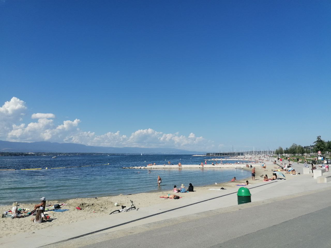 Bathers wade in Lake Geneva beside the sandy curves of Plage des Eaux-Vives and its paved promenade.