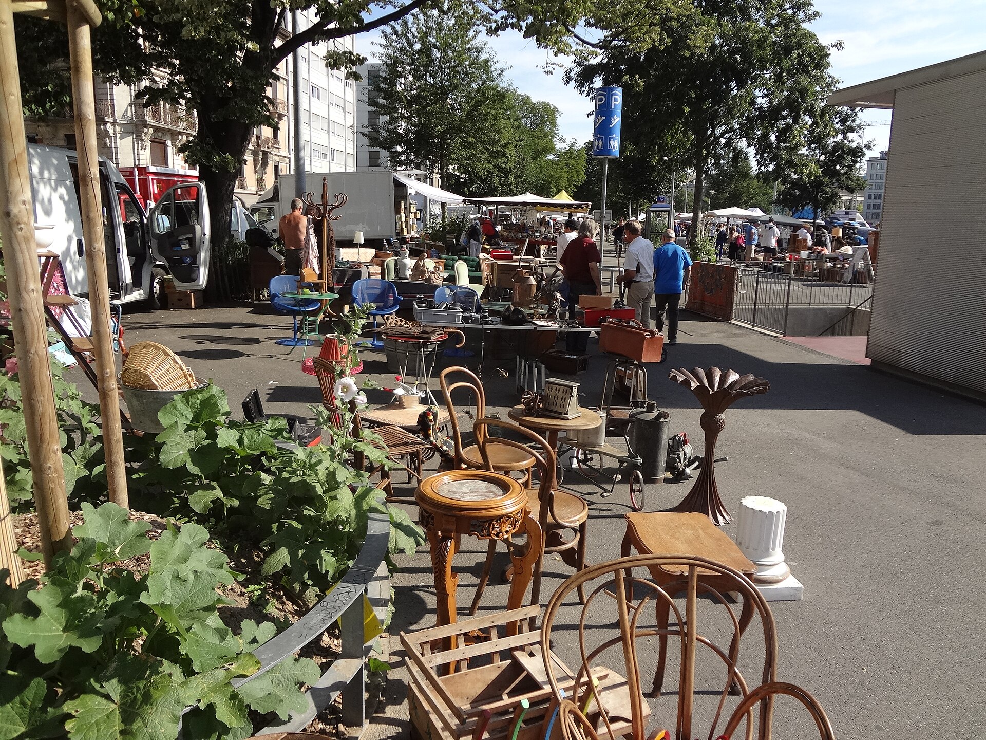 Bentwood chairs, small tables and ornaments stand beside a busy aisle at the Plainpalais flea market in Geneva.