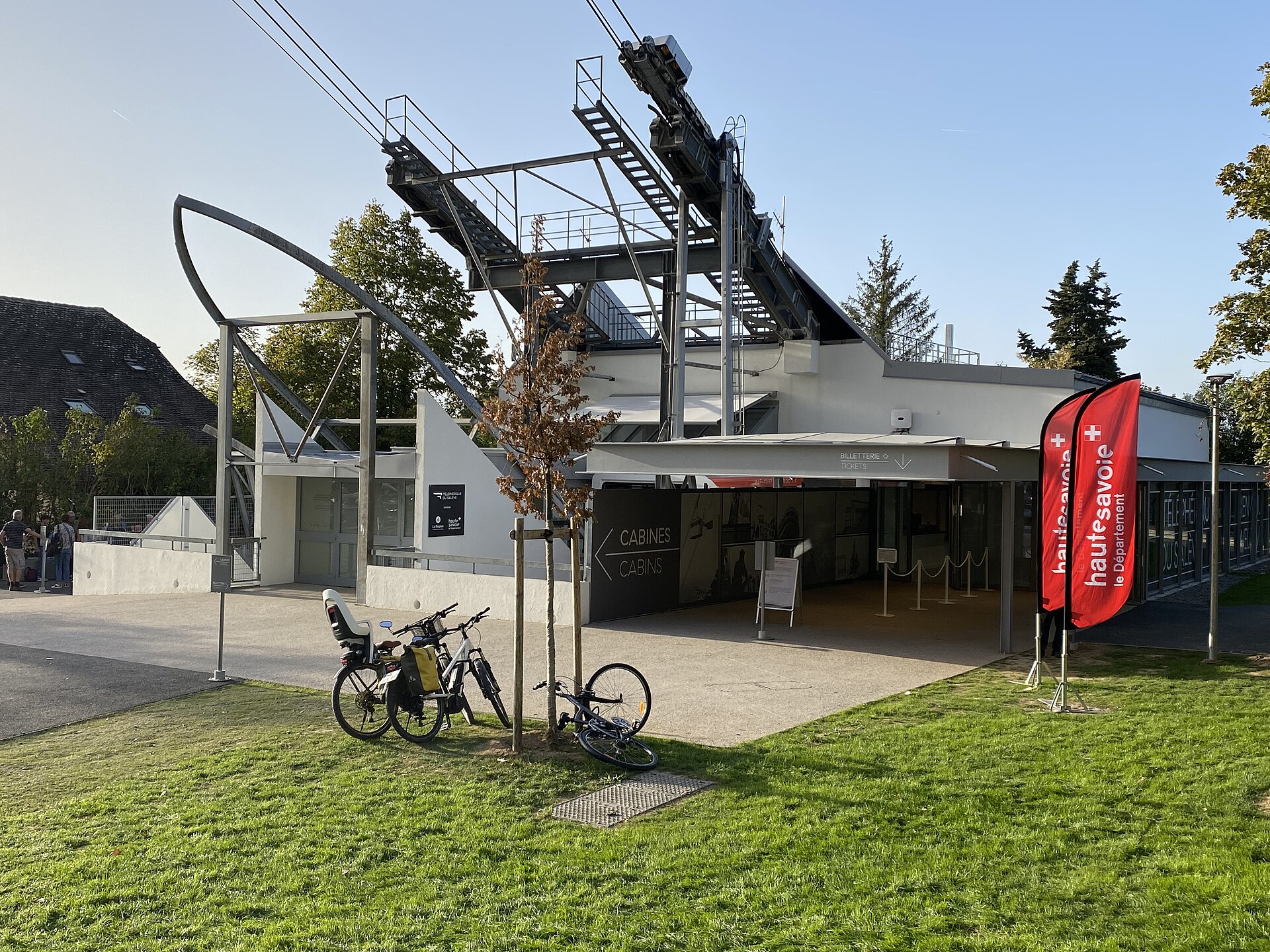 Bicycles stand outside the Salève lower cable-car station beneath steel supports and overhead cables on a sunny day.