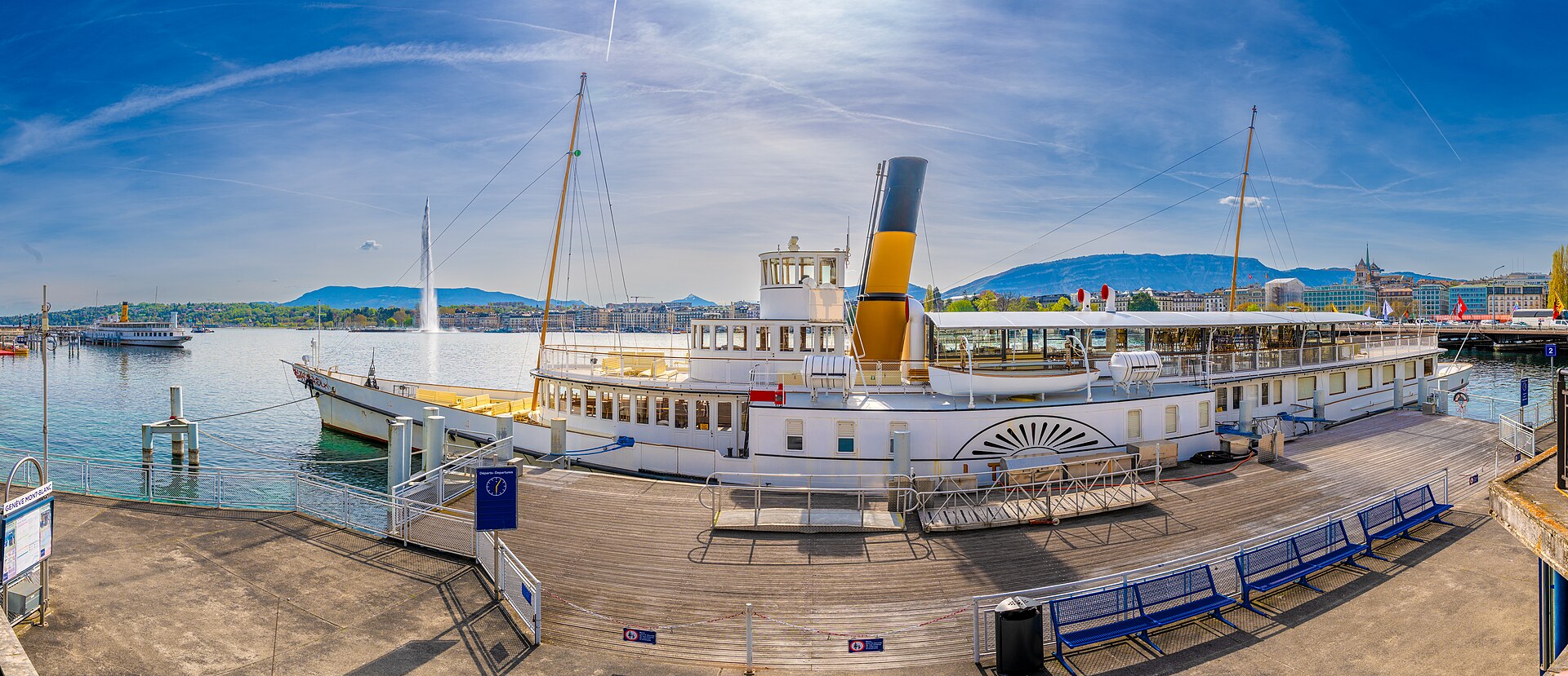 Blue benches line the quay at Geneva's Mont-Blanc landing stage, beside the moored paddle steamer Italie, with the harbour and the Salève beyond.