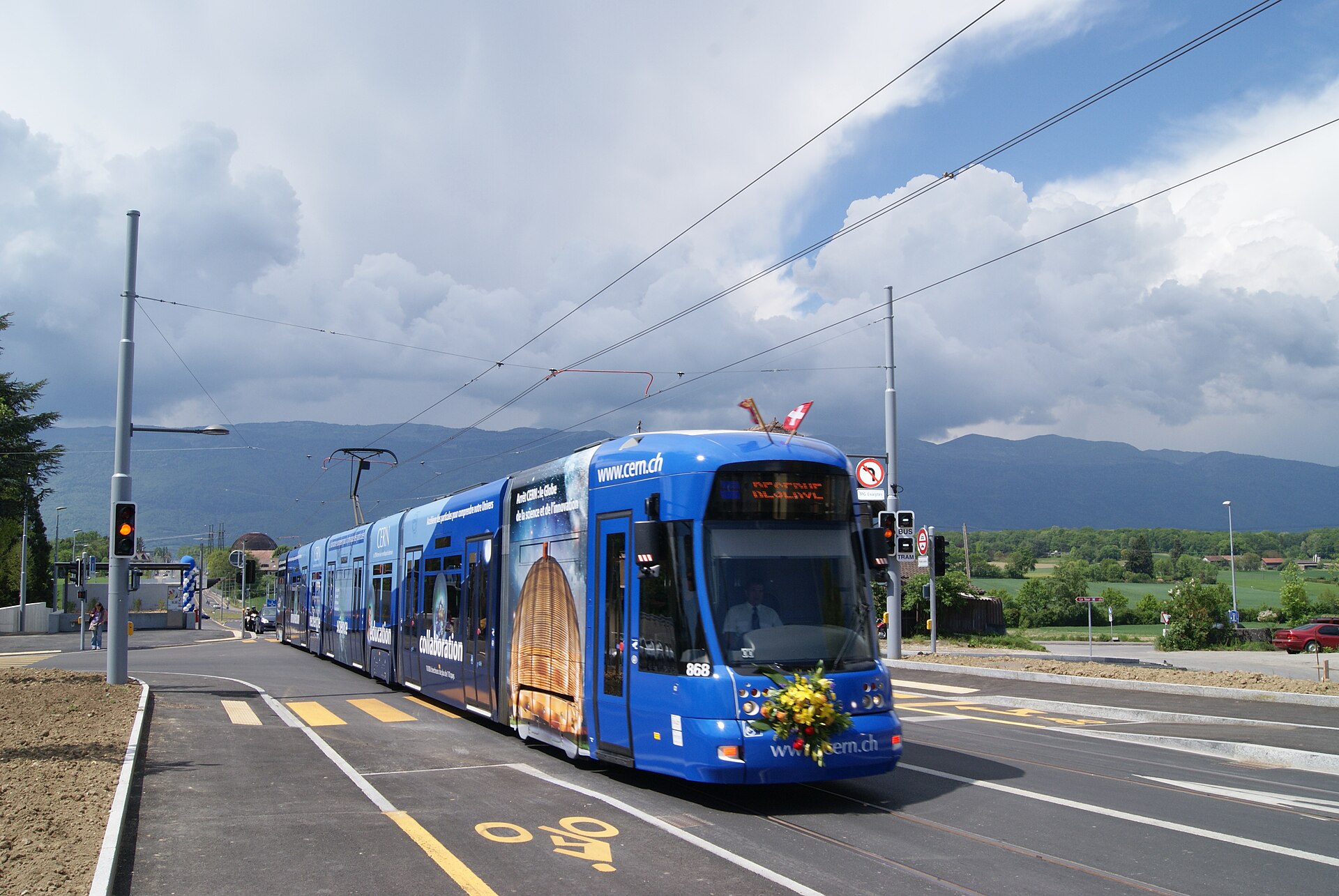 A blue CERN-liveried Geneva tram approaches along Route de Meyrin, with flowers on its front and hills in the background.