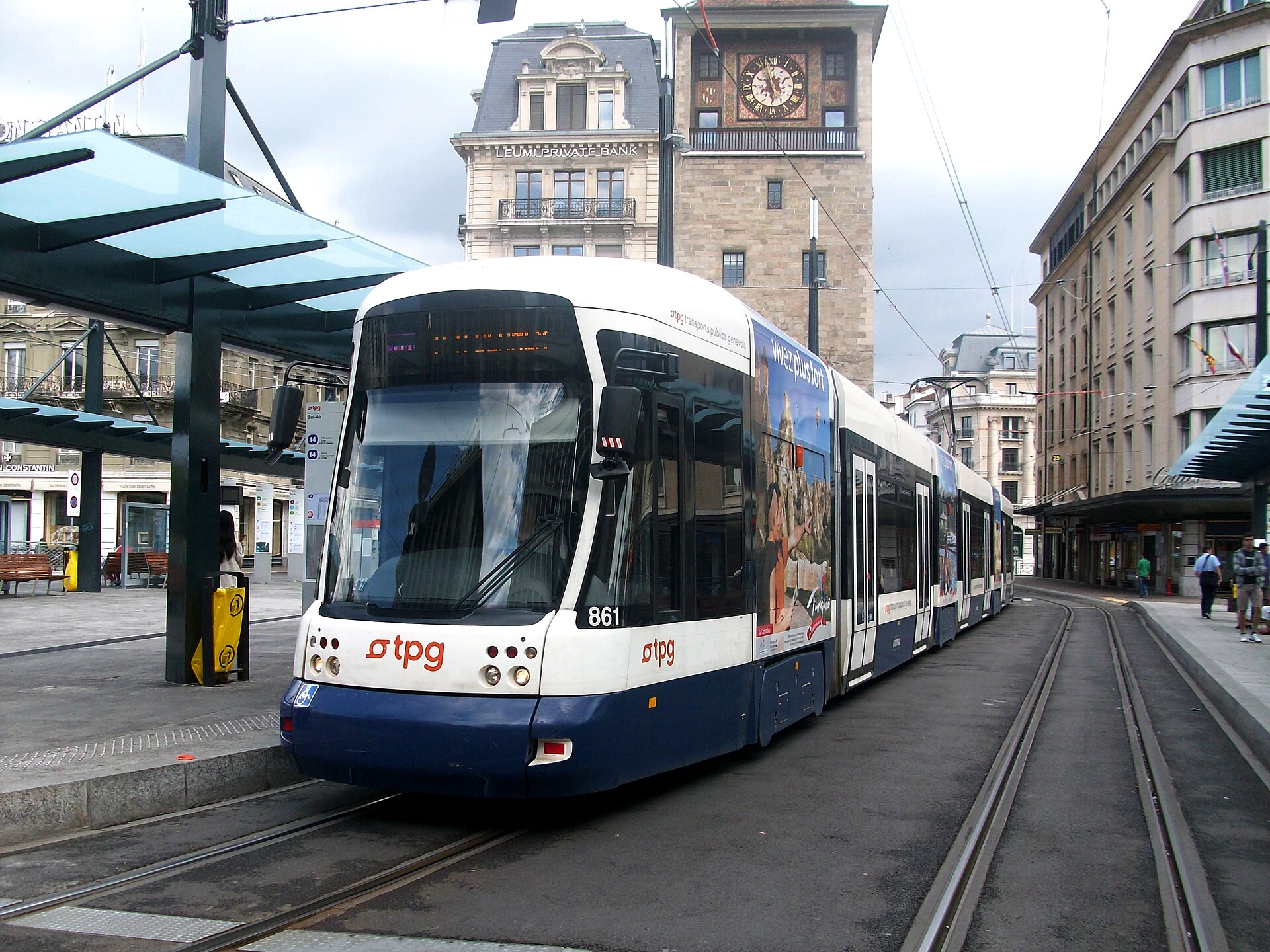 Blue and white TPG tram at Geneva's Bel-Air stop, beside a covered platform with the clock tower behind.
