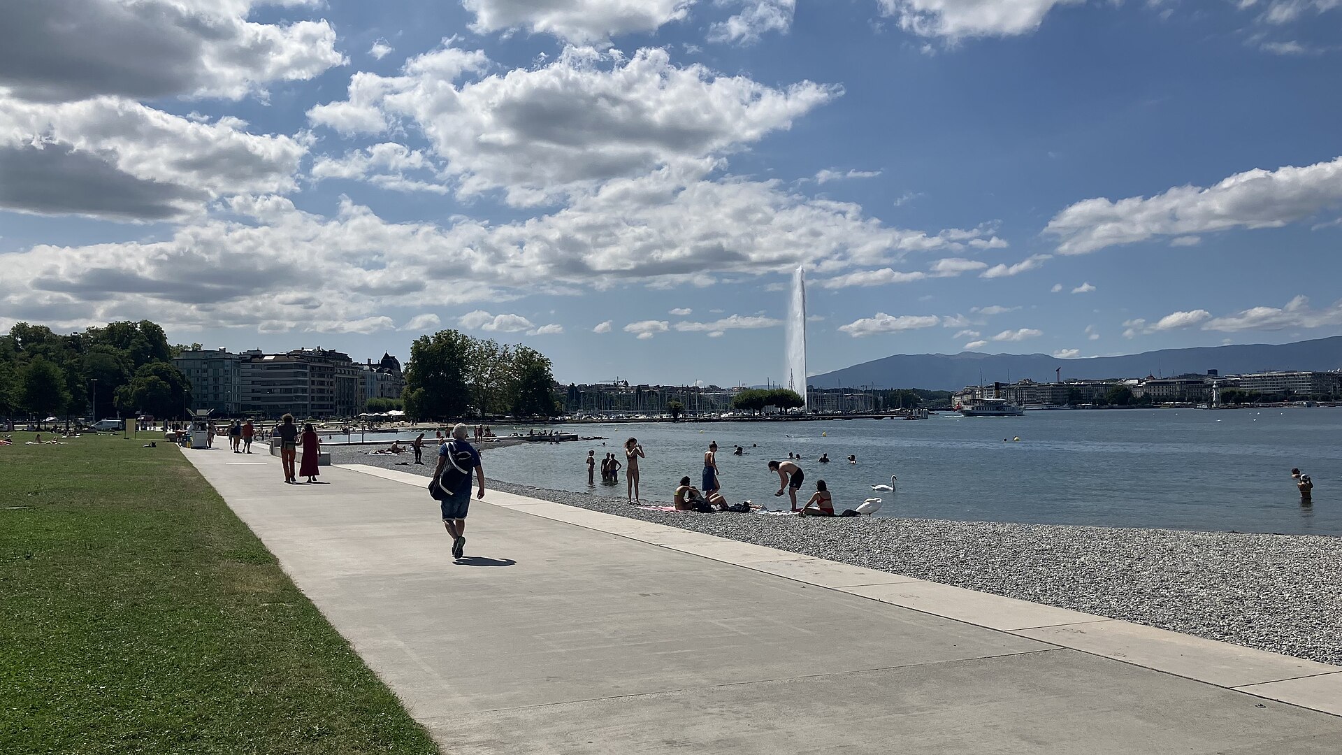 A broad promenade follows Plage des Eaux-Vives beside bathers in Lake Geneva, with the Jet d'Eau beyond.