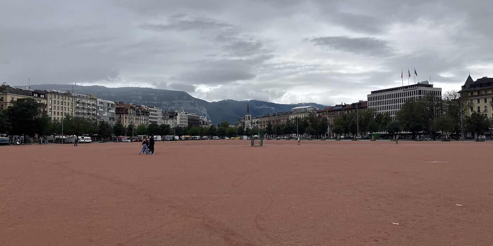 The broad red surface of Geneva's Plaine de Plainpalais stretches toward surrounding buildings, with the Salève ridge beneath a cloudy sky.