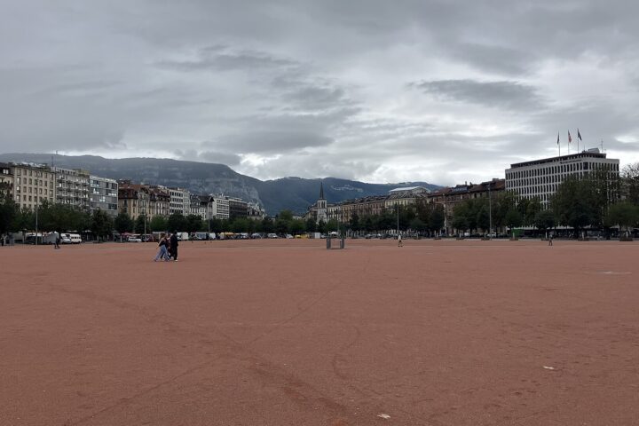 The broad red surface of Geneva's Plaine de Plainpalais stretches toward surrounding buildings, with the Salève ridge beneath a cloudy sky.