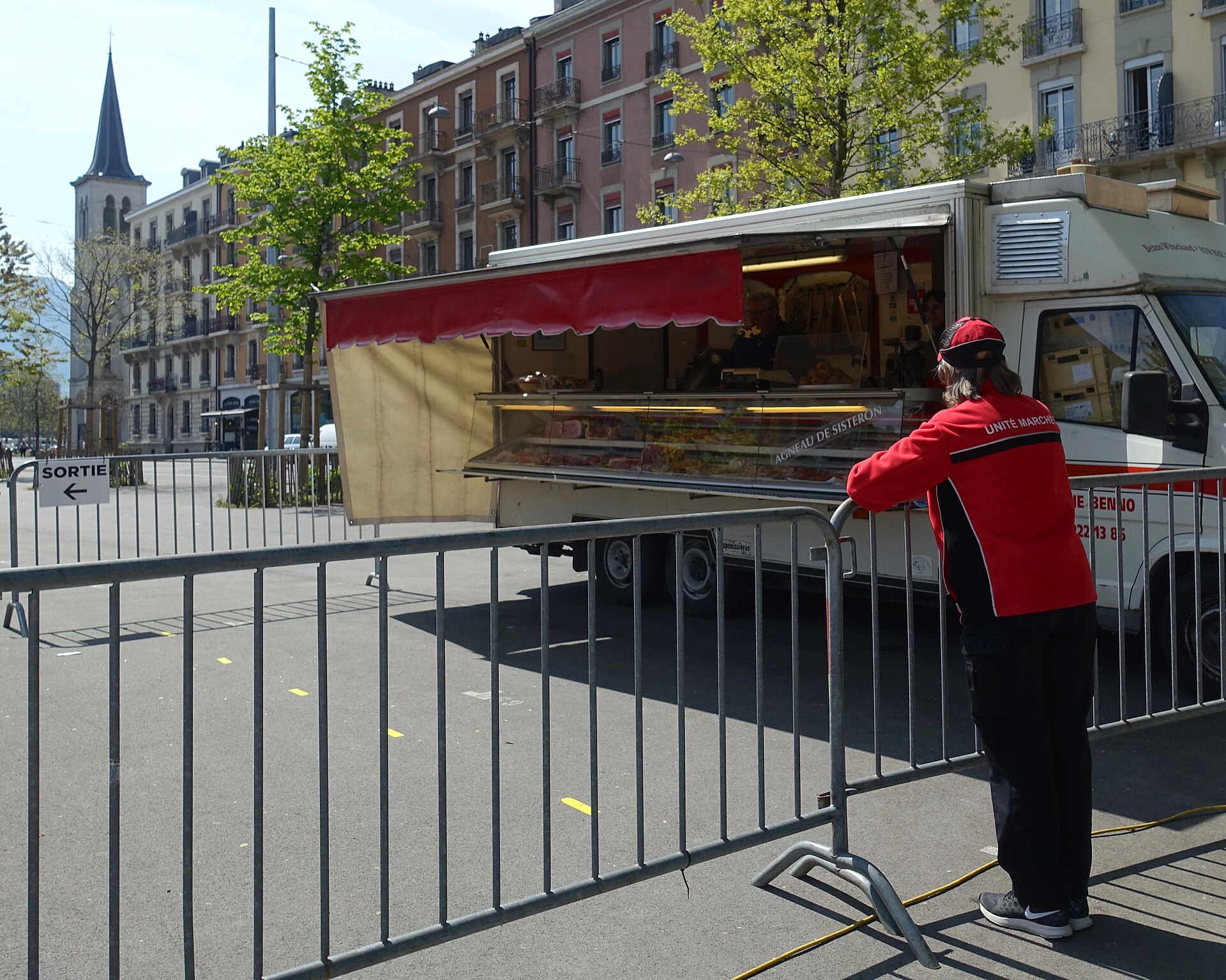 A butcher truck displays meat at Plainpalais market in Geneva while a market worker stands beside temporary barriers.