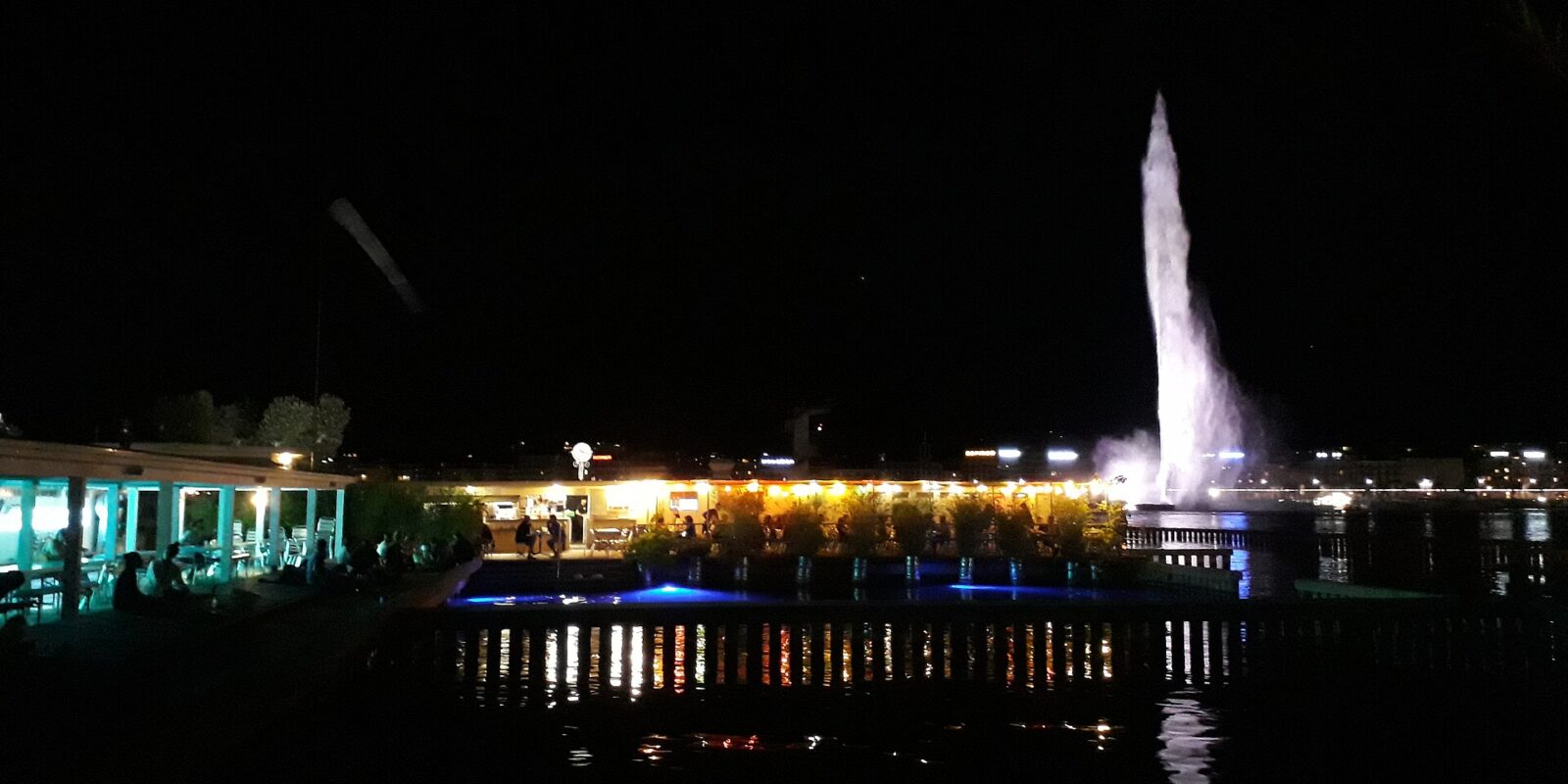 The Buvette des Bains glows beside the lake at night, with diners on the terrace and Geneva's illuminated Jet d'Eau behind.