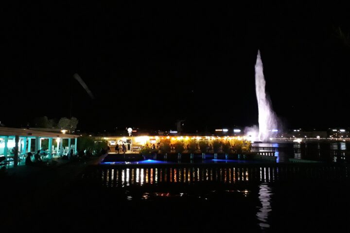 The Buvette des Bains glows beside the lake at night, with diners on the terrace and Geneva's illuminated Jet d'Eau behind.