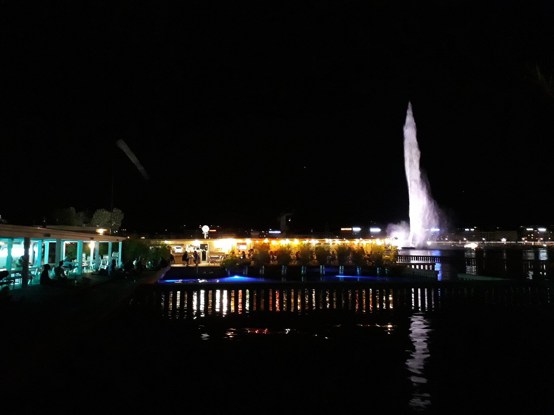 The Buvette des Bains glows beside the lake at night, with diners on the terrace and Geneva's illuminated Jet d'Eau behind.