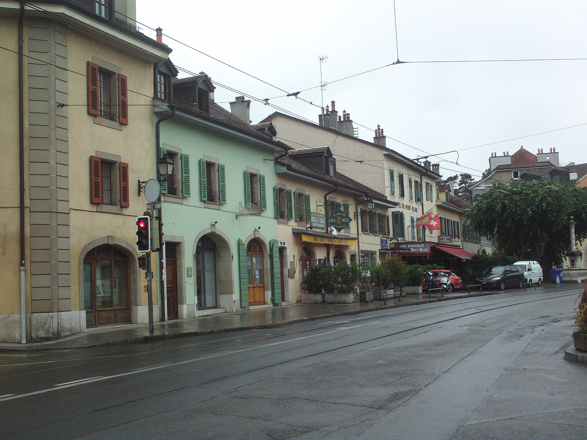 Cafe and restaurant fronts line Rue Ancienne in Carouge, with colourful awnings and terrace planters beneath low shuttered houses.