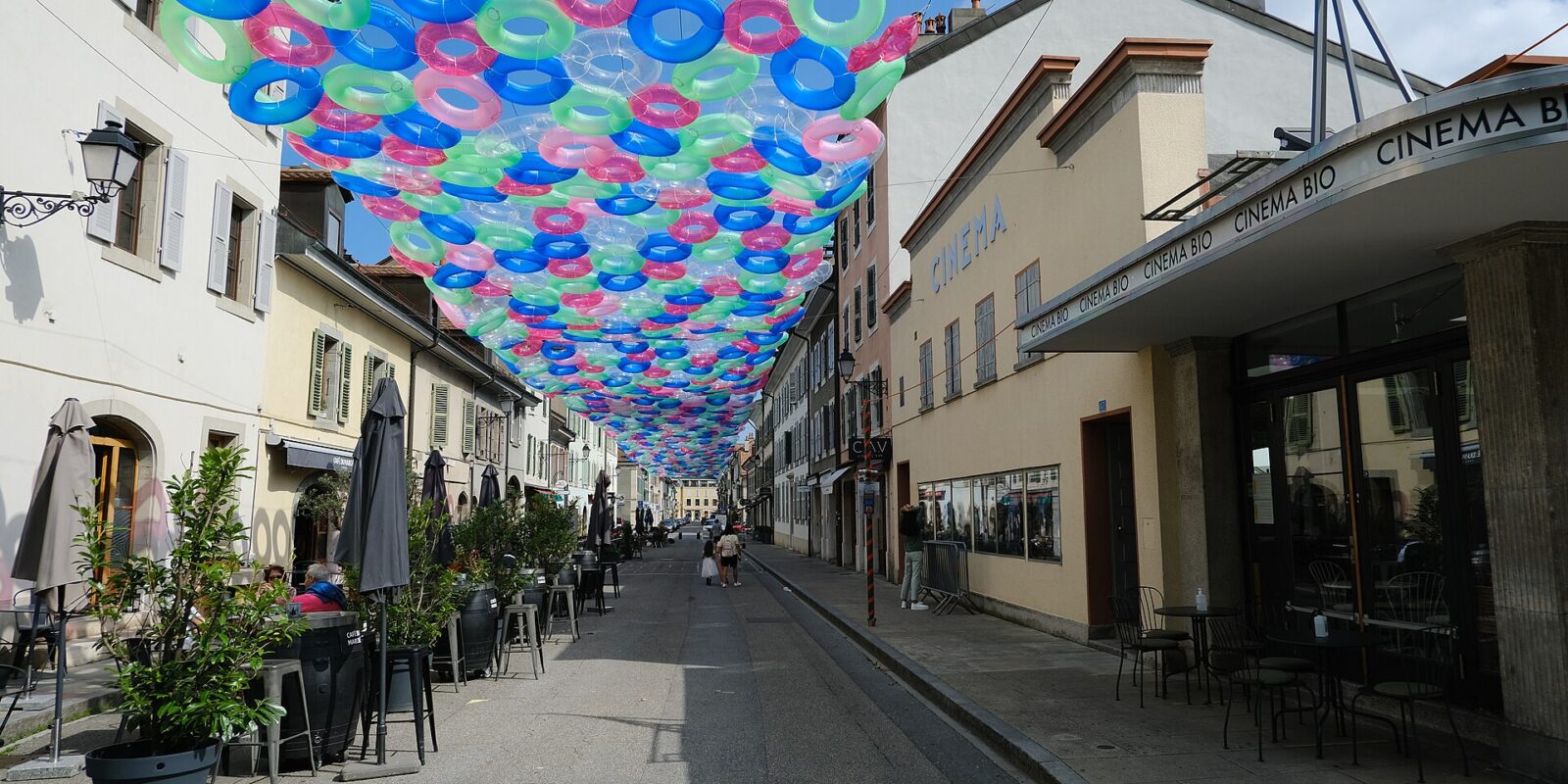 Cafe tables line Rue Saint-Joseph in Carouge beneath colourful hanging rings, beside low shuttered buildings and Cinema Bio.