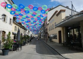 Cafe tables line Rue Saint-Joseph in Carouge beneath colourful hanging rings, beside low shuttered buildings and Cinema Bio.