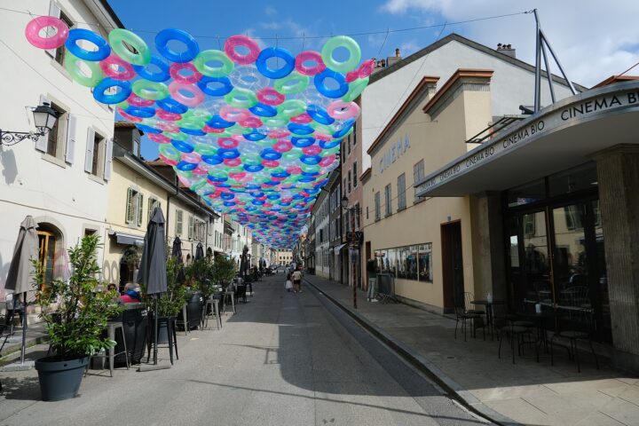 Cafe tables line Rue Saint-Joseph in Carouge beneath colourful hanging rings, beside low shuttered buildings and Cinema Bio.