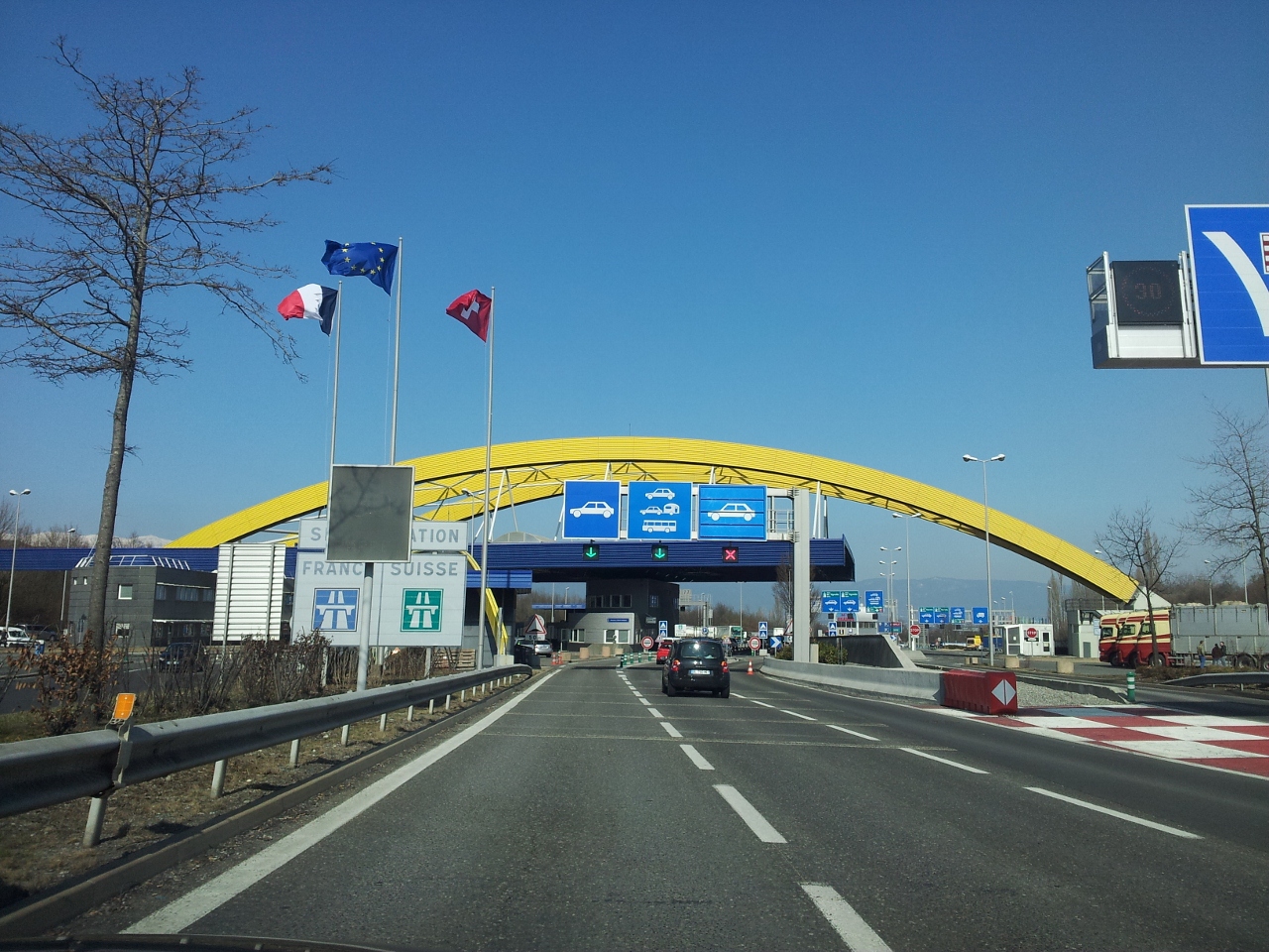 Cars approach the Bardonnex border checkpoint near Geneva beneath a yellow arch beside French, European Union and Swiss flags.