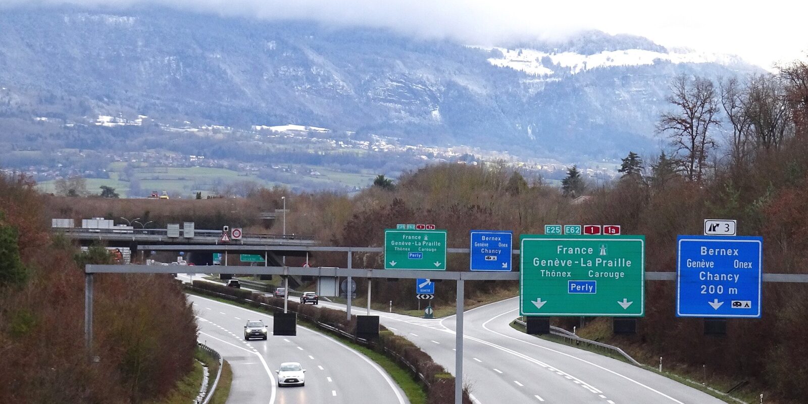 Cars approach the Bernex exit on Geneva's A1 motorway, with green direction signs and the snow-covered Salève behind.